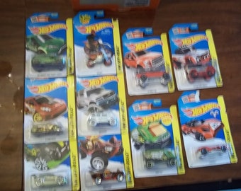 Könnte beinhalten: Eine Sammlung von zehn Hot Wheels Spielzeugautos in ihrer Originalverpackung. Die Autos sind alle in verschiedenen Farben und Stilen, darunter ein grüner LKW, ein roter LKW, ein weißer LKW, ein grüner Monstertruck, ein roter Sportwagen, ein silbernes Auto, ein grünes Auto, ein rotes Auto, ein schwarzes Auto und ein silbernes Auto. Die Verpackung ist blau und gelb mit dem Hot Wheels Logo.