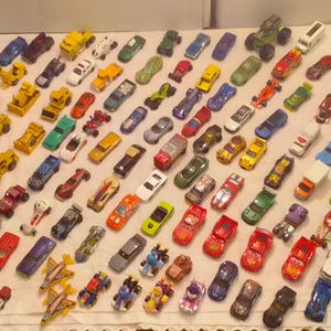 Può includere: Una collezione di modellini di auto, camion e veicoli da cantiere di vari colori, tra cui rosso, giallo, blu e verde. I veicoli in miniatura sono disposti su una superficie bianca, mostrando una vasta gamma di modelli e design.
