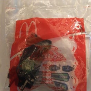 Może przedstawiać: Plastikowa torba zawierająca czarny i zielony samochodzik z nadrukiem "Hot Wheels Raceracers". Torba ma r&oacute;wnież czerwone logo McDonald's i napis "RD-06" nadrukowany na niej.