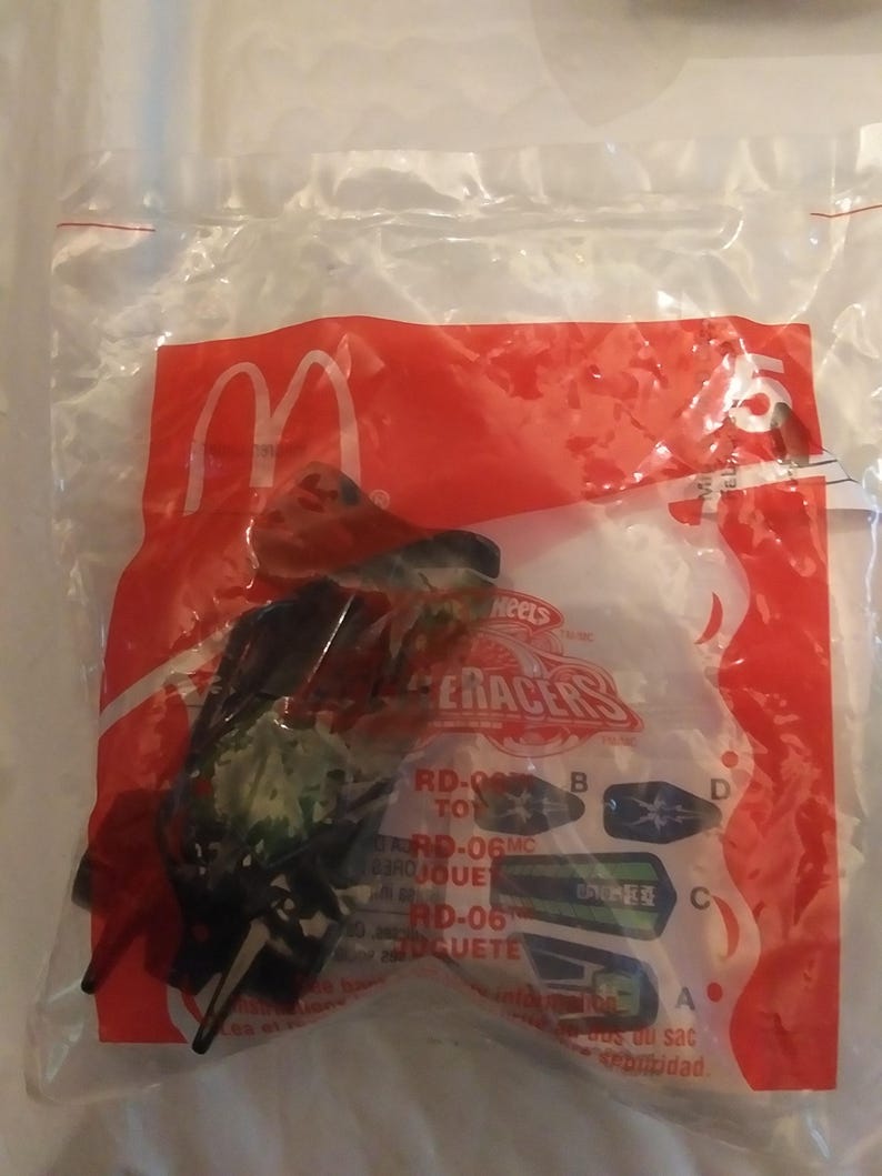 Może przedstawiać: Plastikowa torba zawierająca czarny i zielony samochodzik z nadrukiem "Hot Wheels Raceracers". Torba ma r&oacute;wnież czerwone logo McDonald's i napis "RD-06" nadrukowany na niej.