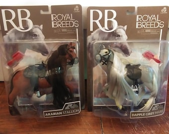 May include: Two toy horse figurines in plastic packaging. The horse on the left is brown with a black mane and tail. The horse on the right is gray with a white mane and tail. Both horses have saddles and bridles. The packaging includes the words "Royal Breeds" and the names of the horses, "Arabian Stallion" and "Dapple Grey Mare".