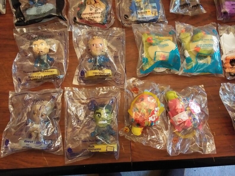 Puede incluir: Una colecci&oacute;n de 10 figuras de juguete de pl&aacute;stico en envases de pl&aacute;stico transparente. Las figuras incluyen personajes de pel&iacute;culas y programas de televisi&oacute;n populares, como Thor, Hulk y personajes de la pel&iacute;cula de Lego. El envase es morado y presenta el nombre del personaje y una peque&ntilde;a imagen del personaje.