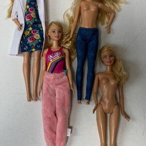 May include: Four blonde Barbie dolls with different outfits. One doll is wearing a white lab coat over a floral dress, another is wearing a pink fuzzy pajama top and pants, another is wearing a blue tank top with the word "Malibu" on it and blue jeans, and the last doll is nude.