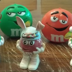 May include: Five M&M's character figurines. A green M&M with antlers, a green M&M wearing a Santa hat, a red M&M with bunny ears, a green M&M with a large M on its chest, and a red M&M with a large M on its chest.