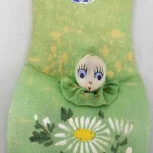 May include: A green felt pincushion with a painted daisy and a small doll with blue eyes peeking out. The pincushion has a blue and white circular sticker with the text "1933 A Century of Progress Chicago".