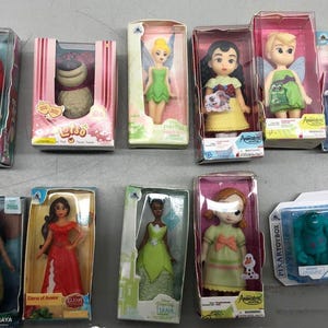 K&ouml;nnte beinhalten: Eine Sammlung von Disney-Charakterpuppen in ihrer Originalverpackung. Die Puppen umfassen Arielle, Lilo, Tinkerbell, Cinderella und Tiana. Die Puppen sind in verschiedenen Outfits und Posen, mit leuchtenden Farben und detaillierten Designs. Die Sammlung enth&auml;lt auch eine Sully-Figur.