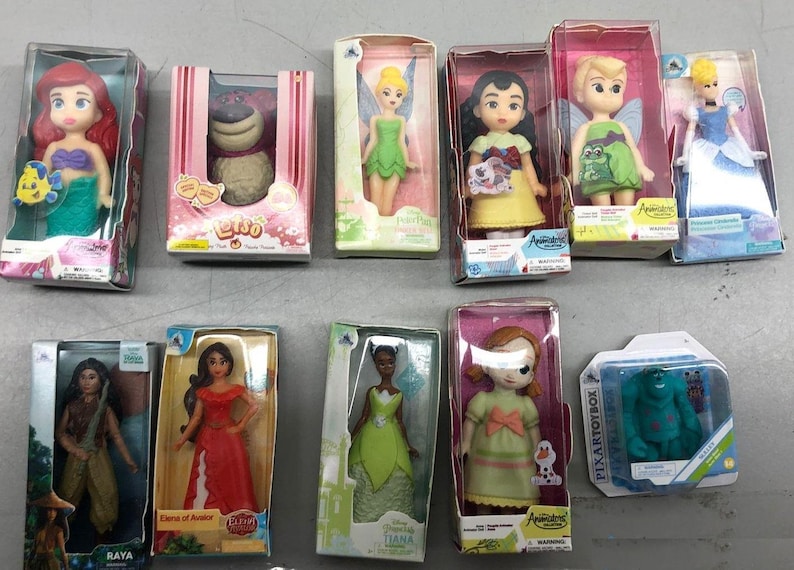 K&ouml;nnte beinhalten: Eine Sammlung von Disney-Charakterpuppen in ihrer Originalverpackung. Die Puppen umfassen Arielle, Lilo, Tinkerbell, Cinderella und Tiana. Die Puppen sind in verschiedenen Outfits und Posen, mit leuchtenden Farben und detaillierten Designs. Die Sammlung enth&auml;lt auch eine Sully-Figur.