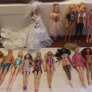 Puede incluir: Una colección de 18 muñecas, algunas con vestidos, otras con trajes de baño y otras con ropa informal. Las muñecas están dispuestas en dos filas sobre una superficie blanca.