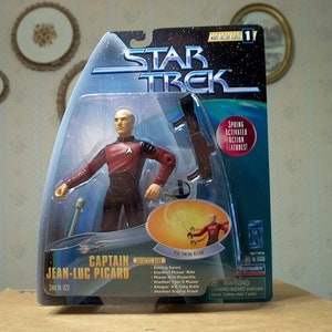 Może przedstawiać: Figurka akcji Star Trek kapitana Jeana-Luca Picarda w jego czerwonym mundurze. Figurka ma funkcje akcji uruchamiane sprężyną i jest wyposażona w miecz i fazer. Opakowanie ma niebieskie i białe tło z logo Star Trek.