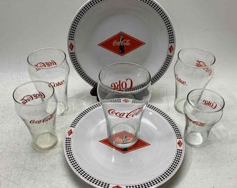 May include: A set of Coca-Cola themed glassware, including a large serving bowl, four glasses, and a smaller serving plate. The bowl and plate feature a black and white checkered pattern with a red Coca-Cola logo.