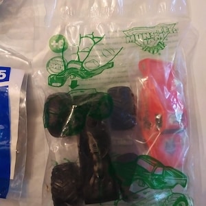 Może przedstawiać: Przezroczysty plastikowy worek zawierający ciężar&oacute;wkę Monster Jam. Ciężar&oacute;wka jest zielona i czarna z czerwonym logo Monster Jam. Worek zawiera r&oacute;wnież mniejszy czerwony samoch&oacute;d. Worek ma logo Burger King i napis "Monster Jam".