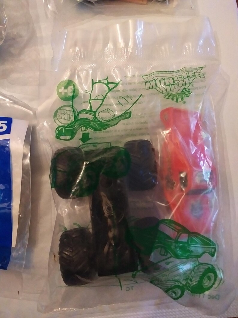 Może przedstawiać: Przezroczysty plastikowy worek zawierający ciężar&oacute;wkę Monster Jam. Ciężar&oacute;wka jest zielona i czarna z czerwonym logo Monster Jam. Worek zawiera r&oacute;wnież mniejszy czerwony samoch&oacute;d. Worek ma logo Burger King i napis "Monster Jam".