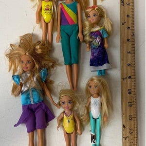 May include: Seven Barbie dolls in various outfits. The dolls have blonde hair and are wearing colorful clothing. The dolls are arranged in a row on a wooden surface. The dolls are all different sizes and have different facial expressions.