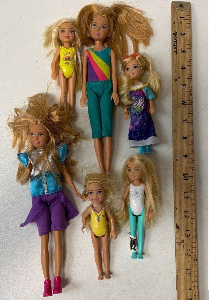 May include: Seven Barbie dolls in various outfits. The dolls have blonde hair and are wearing colorful clothing. The dolls are arranged in a row on a wooden surface. The dolls are all different sizes and have different facial expressions.