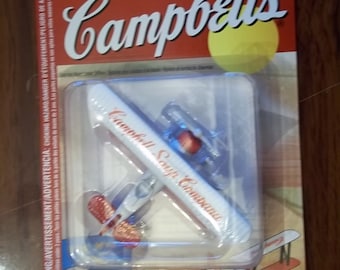May include: A red, white, and blue toy airplane with the words "Campbell Soup Company" printed on the side. The airplane is packaged in a cardboard box with the Campbell's Soup logo.
