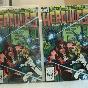 以下が含まれることがあります： 「Hercules, Prince of Power」というタイトルの2冊のコミック。表紙には、筋肉質な男性と白い衣装の女性が描かれており、Marvel Comicsのロゴがあります。背景には「100,000 REWARD」の文字が含まれています。