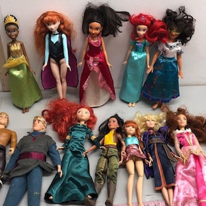 May include: A collection of dolls featuring various characters in colorful outfits. The dolls have different hair colors and styles, and wear dresses, gowns, and other costumes. Some dolls have accessories like crowns.