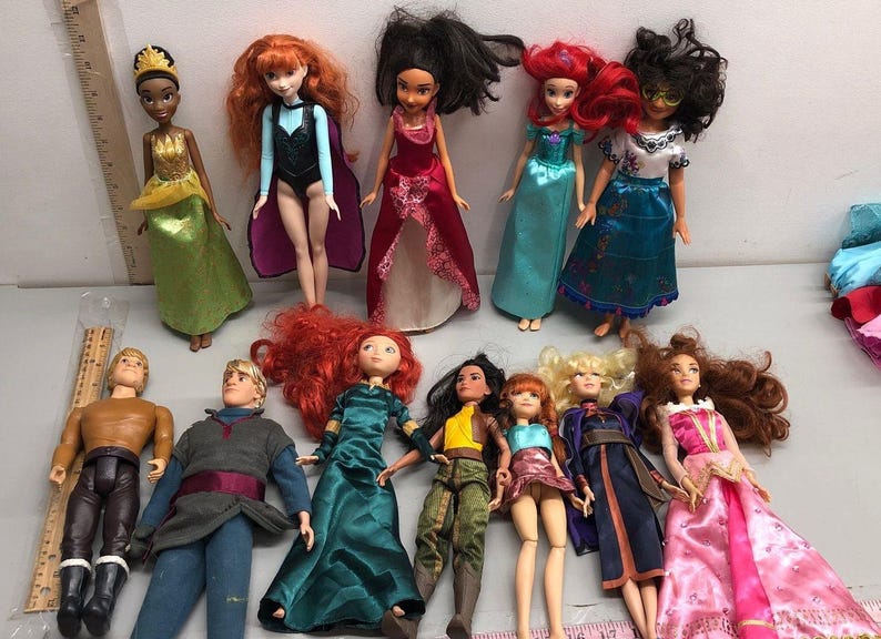 May include: A collection of dolls featuring various characters in colorful outfits. The dolls have different hair colors and styles, and wear dresses, gowns, and other costumes. Some dolls have accessories like crowns.