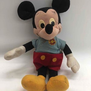 Puede incluir: Un juguete de peluche vintage de Mickey Mouse con orejas negras, una camisa azul con una flor amarilla, pantalones cortos rojos con botones amarillos y zapatos negros.