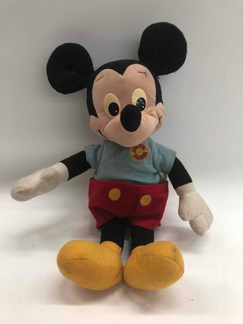 Puede incluir: Un juguete de peluche vintage de Mickey Mouse con orejas negras, una camisa azul con una flor amarilla, pantalones cortos rojos con botones amarillos y zapatos negros.