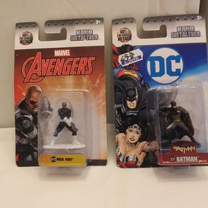 May include: Two Nano Metalfigs action figures in their original packaging. One features Nick Fury from Marvel's Avengers, and the other features Batman from DC Comics. The packaging is red and orange for Avengers and blue for Batman.