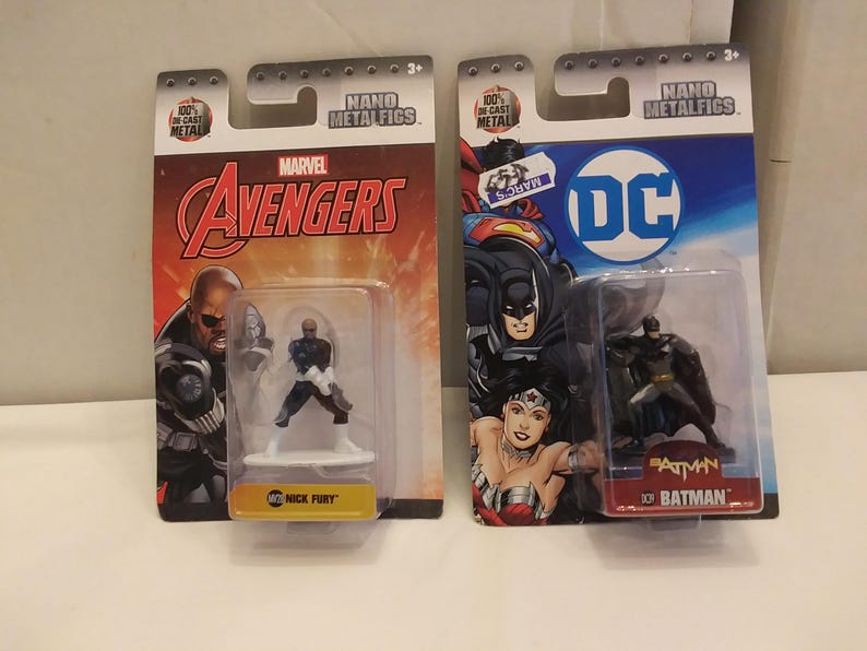 May include: Two Nano Metalfigs action figures in their original packaging. One features Nick Fury from Marvel's Avengers, and the other features Batman from DC Comics. The packaging is red and orange for Avengers and blue for Batman.