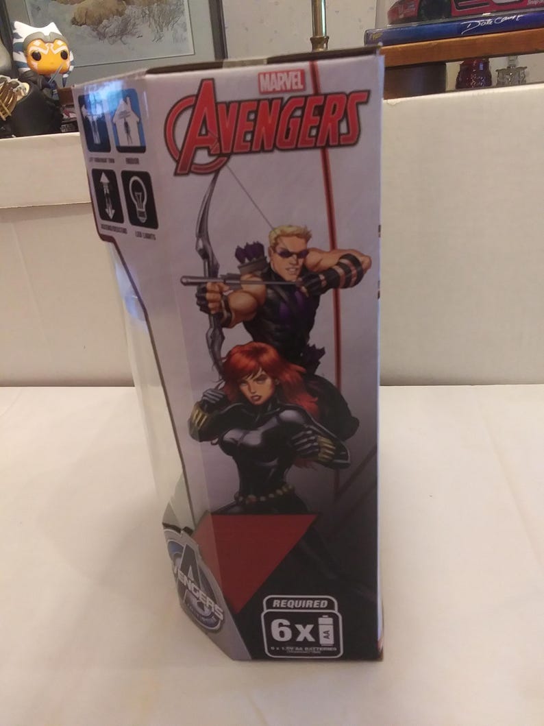 May include: A cardboard box for a Marvel Avengers toy. The box features a red, white and black design with images of Hawkeye and Black Widow. The text "MARVEL AVENGERS" is printed in large, bold letters on the box. The box also includes the text "REQUIRED 6x AA BATTERIES".