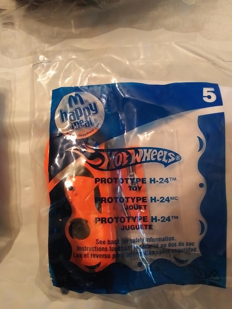 Może przedstawiać: Plastikowa torba zawierająca czerwony samochodzik Hot Wheels. Torba jest niebieska i pomarańczowa i ma nadrukowane słowa "Hot Wheels", "Prototype H-24" i "Happy Meal".