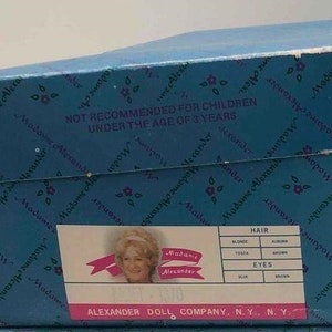 May include: Blue and white box with the text "Madame Alexander" and "NOT RECOMMENDED FOR CHILDREN UNDER THE AGE OF 3 YEARS". The box also has a picture of a doll with blonde hair and blue eyes. The text "ALEXANDER DOLL COMPANY, N.Y., N.Y." is at the bottom of the box.
