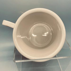 Puede incluir: Una taza de cer&aacute;mica blanca con asa, vista desde arriba. El interior de la taza es de color crema claro. La taza est&aacute; sobre un expositor transparente inclinado sobre un fondo azul claro.