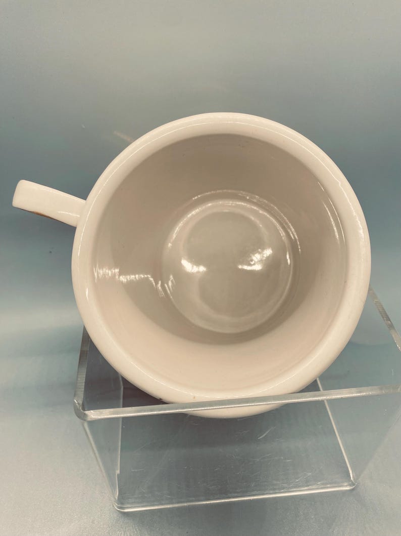 Puede incluir: Una taza de cer&aacute;mica blanca con asa, vista desde arriba. El interior de la taza es de color crema claro. La taza est&aacute; sobre un expositor transparente inclinado sobre un fondo azul claro.