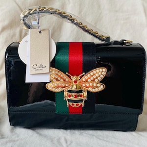 Calin: Bolso vegano negro con forma de abeja y correa cruzada con billetera a juego. Nuevo en su tipo. Sin abrir. Cadena para llevar en la mano y asa o bandolera de cuero vegano.