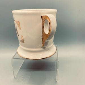 Puede incluir: Una taza de afeitar de cer&aacute;mica blanca con asa y ribete dorados. La taza tiene letras doradas en el lateral y se apoya en un soporte de acr&iacute;lico transparente. La taza tiene un dise&ntilde;o cl&aacute;sico y vintage.