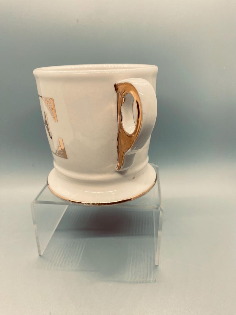 Puede incluir: Una taza de afeitar de cer&aacute;mica blanca con asa y ribete dorados. La taza tiene letras doradas en el lateral y se apoya en un soporte de acr&iacute;lico transparente. La taza tiene un dise&ntilde;o cl&aacute;sico y vintage.