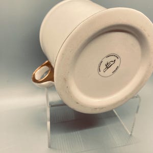 Puede incluir: Taza de cer&aacute;mica blanca con asa y borde dorados. La base de la taza tiene un sello circular negro con un dise&ntilde;o de p&aacute;jaro y el texto "FABRIQUE EN CHINE". La taza est&aacute; sobre un soporte acr&iacute;lico transparente.