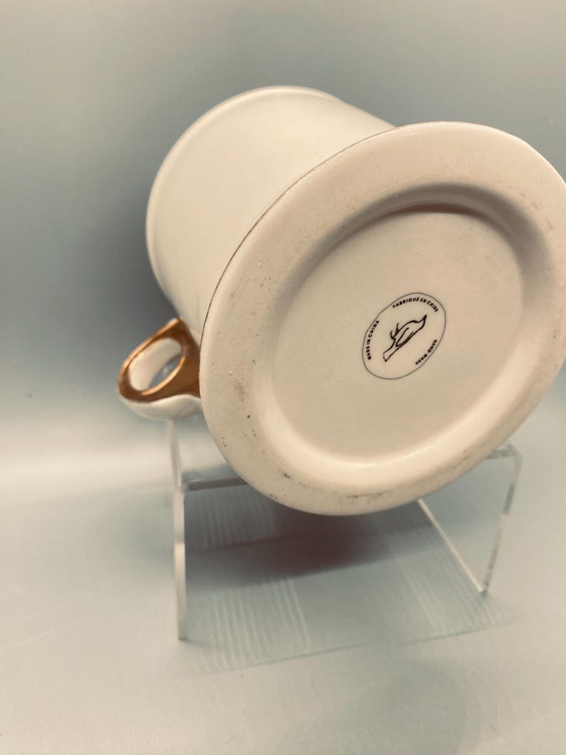 Puede incluir: Taza de cer&aacute;mica blanca con asa y borde dorados. La base de la taza tiene un sello circular negro con un dise&ntilde;o de p&aacute;jaro y el texto "FABRIQUE EN CHINE". La taza est&aacute; sobre un soporte acr&iacute;lico transparente.