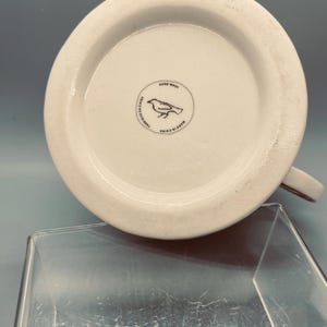 Puede incluir: Taza de cer&aacute;mica blanca con asa marr&oacute;n, vista desde abajo. La base presenta un logotipo circular con una ilustraci&oacute;n de un p&aacute;jaro y las palabras "Hand Wash" y "Made in China".