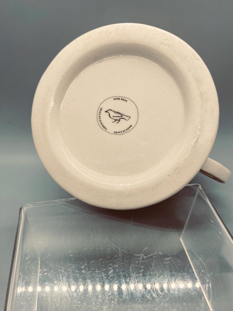 Puede incluir: Taza de cer&aacute;mica blanca con asa marr&oacute;n, vista desde abajo. La base presenta un logotipo circular con una ilustraci&oacute;n de un p&aacute;jaro y las palabras "Hand Wash" y "Made in China".