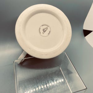 Puede incluir: Taza de cer&aacute;mica blanca con interior marr&oacute;n, vista desde abajo. La base presenta un emblema circular con texto y un dise&ntilde;o dibujado a mano. La taza est&aacute; colocada sobre un expositor de acr&iacute;lico transparente, con un fondo azul claro.