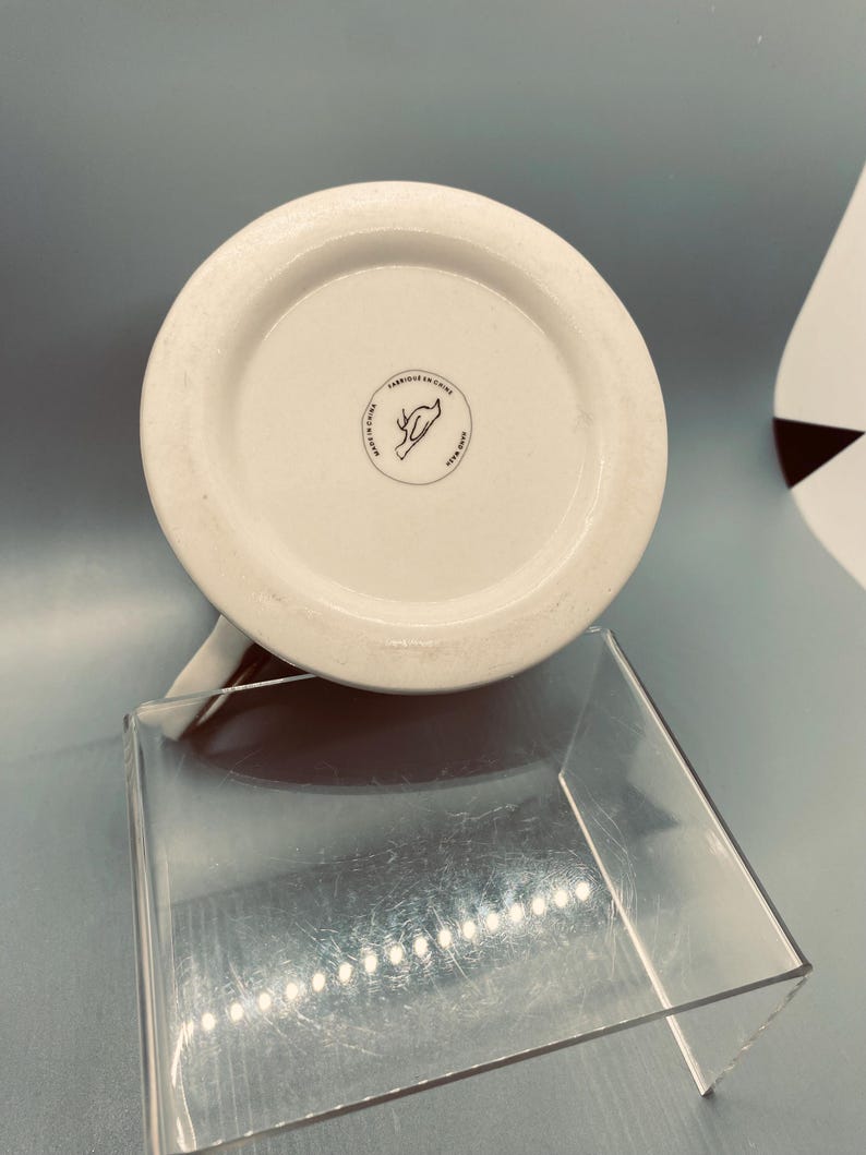 Puede incluir: Taza de cer&aacute;mica blanca con interior marr&oacute;n, vista desde abajo. La base presenta un emblema circular con texto y un dise&ntilde;o dibujado a mano. La taza est&aacute; colocada sobre un expositor de acr&iacute;lico transparente, con un fondo azul claro.