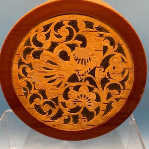 May include: A round wooden box with a carved design of a bird and floral patterns. The box is made of birch wood and has a dark brown stain.