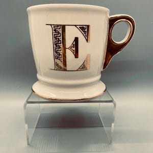 Puede incluir: Taza de caf&eacute; de cer&aacute;mica blanca con asa y ribetes dorados. La letra "E" se muestra en dorado decorativo en la parte delantera. La taza tiene una base de pedestal. Un art&iacute;culo decorativo para la cocina.