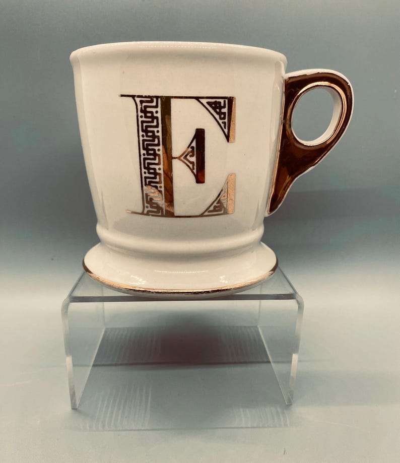 Puede incluir: Taza de caf&eacute; de cer&aacute;mica blanca con asa y ribetes dorados. La letra "E" se muestra en dorado decorativo en la parte delantera. La taza tiene una base de pedestal. Un art&iacute;culo decorativo para la cocina.
