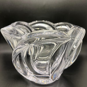 May include: A clear, decorative glass bowl with a unique, wavy design. The bowl has a series of curved edges that create a flower-like shape. The glass is transparent, allowing light to pass through and reflect off the curves.