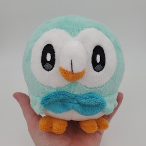 Puede incluir: Un juguete de peluche de un pingüino azul y blanco con una pajarita azul. El pingüino tiene un cuerpo redondo y un pequeño pico. El juguete está hecho de un material suave y mullido.