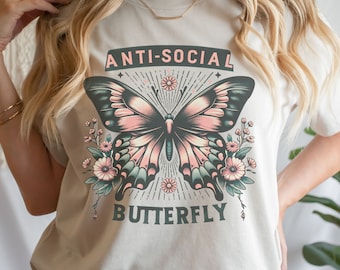May include: Cream-colored t-shirt featuring a butterfly design with the text "ANTI-SOCIAL BUTTERFLY." The butterfly has pink, teal, and black wings, surrounded by floral accents. The text is in a dark green font.