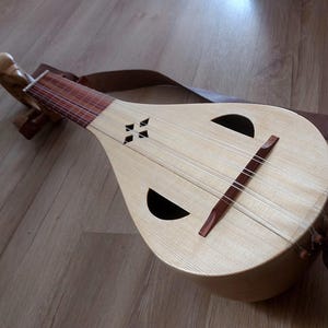 May include: A light-colored, pear-shaped stringed instrument with a brown leather strap. The instrument has two large, crescent-shaped sound holes and a dark brown bridge. The wooden body is light-colored, and the instrument rests on a wooden surface.