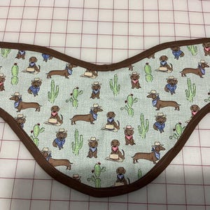 May include: A light blue pet bandana with a brown border. The bandana features a repeating pattern of dachshunds wearing cowboy hats and bandanas, along with green cactus illustrations.