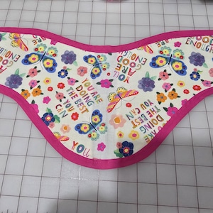 May include: A white bib with a pink trim, decorated with colorful butterflies and flowers. The bib features the text "YOU ARE DOING THE BEST YOU CAN" and "YOU ARE GOOD ENOUGH".
