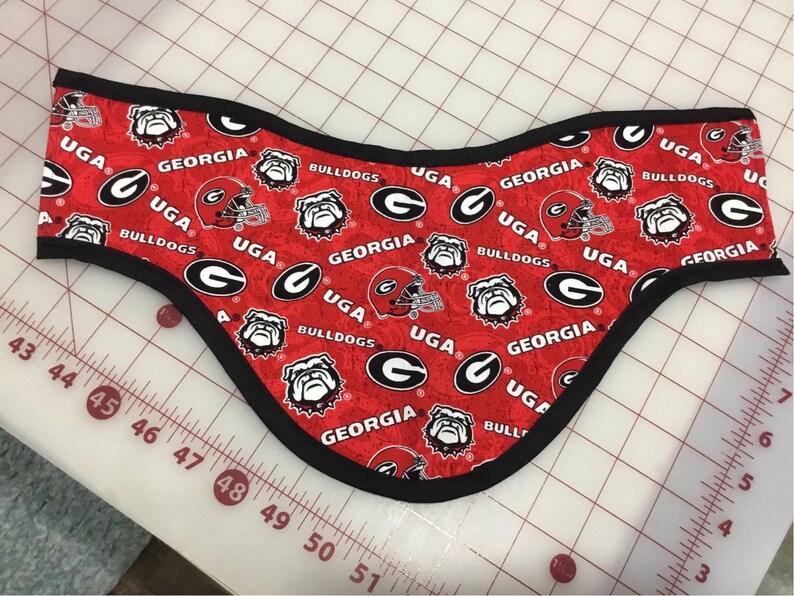 May include: Red fabric neck warmer with a black trim. The fabric has a repeating pattern of the University of Georgia logo, a bulldog mascot, and the words "UGA", "Georgia", and "Bulldogs".